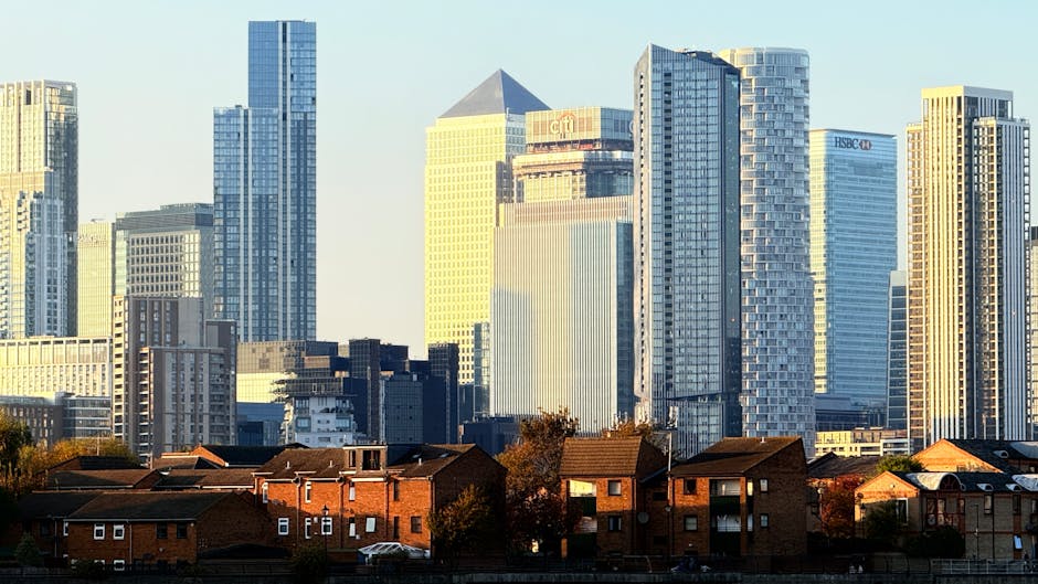 A cityscape featuring a row of low-rise brick residential houses and apartment buildings in the foreground, with a modern skyline of tall office skyscrapers made of glass and steel in the background. 