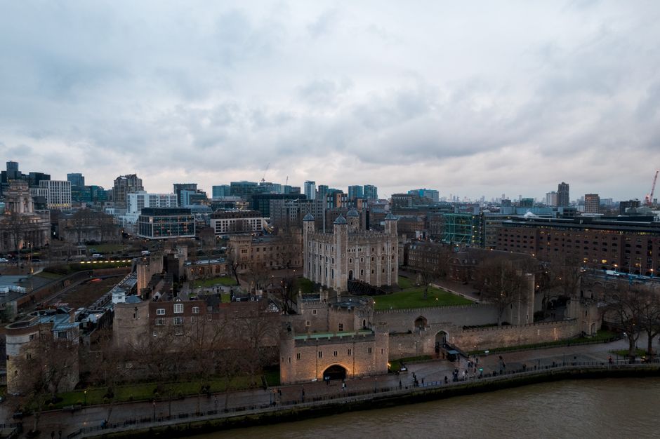 View of the Tower of London during daytime, with a stone fortress featuring four turrets and crenellated walls, partially obscured by green-leaved trees in the foreground. Behind the historic castle,